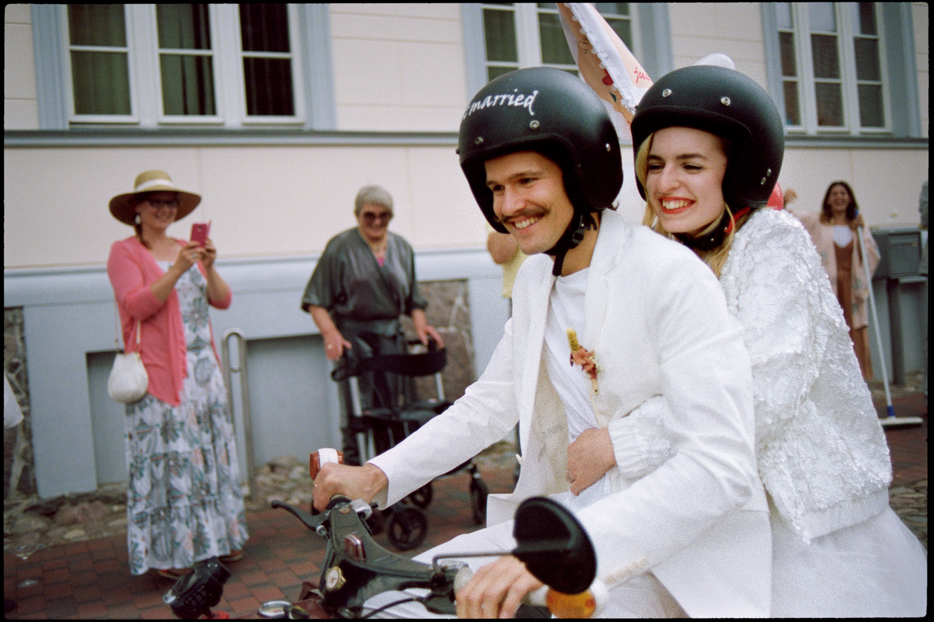 The couple on the scooter, guests celebrating around them, Maike & Daniel wedding in Brandenburg