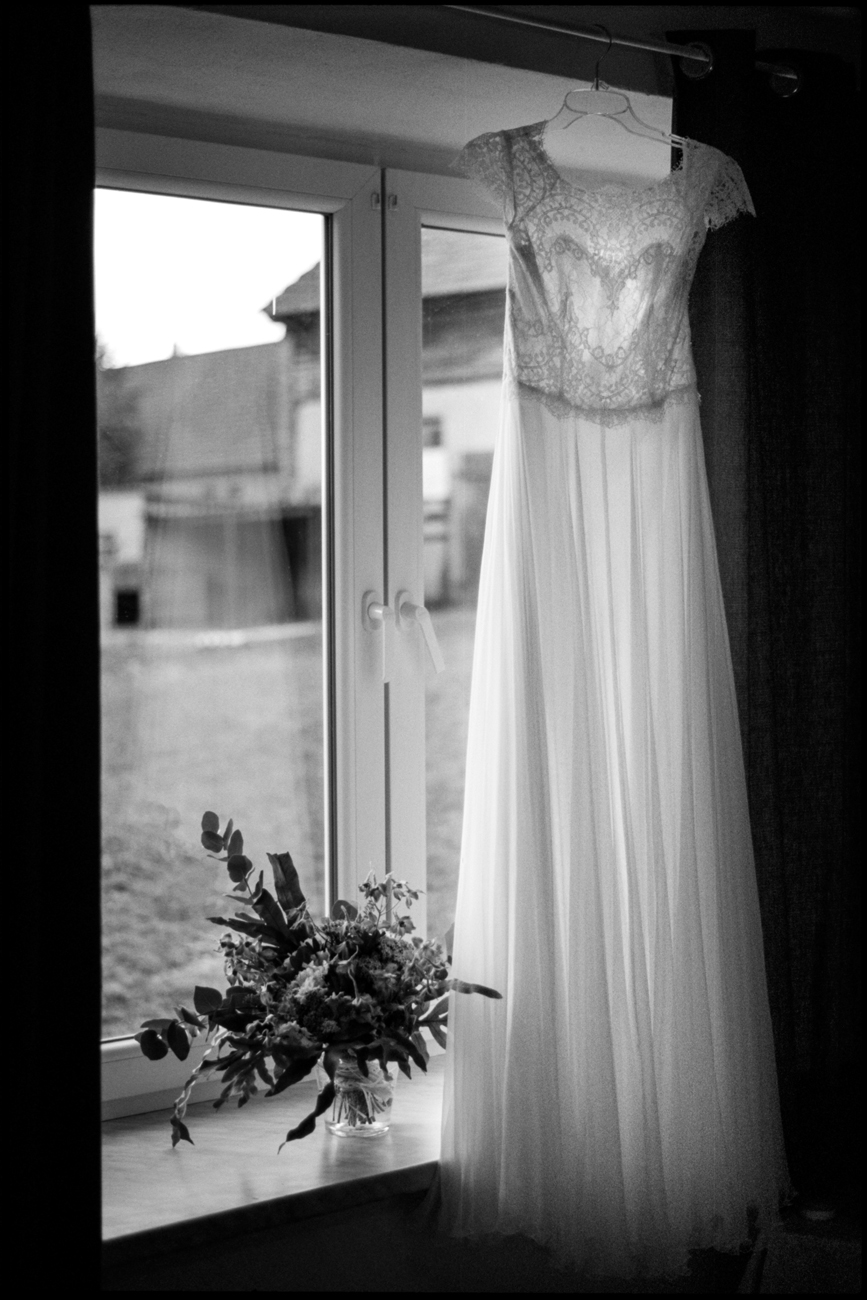 The wedding dress hanging in a window with a bouquet, in black and white, Maike & Daniel wedding in Brandenburg