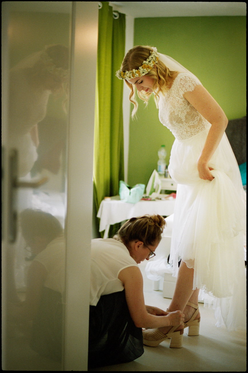Bride in a flower crown having her shoes fastened while getting ready, analogue film photograph by Martina Manz