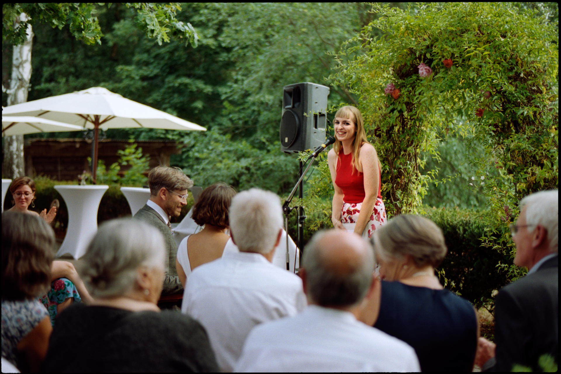 A guest giving a reading to seated guests in the garden, Suelyn & Pascal wedding in Groß Glienicke, Potsdam