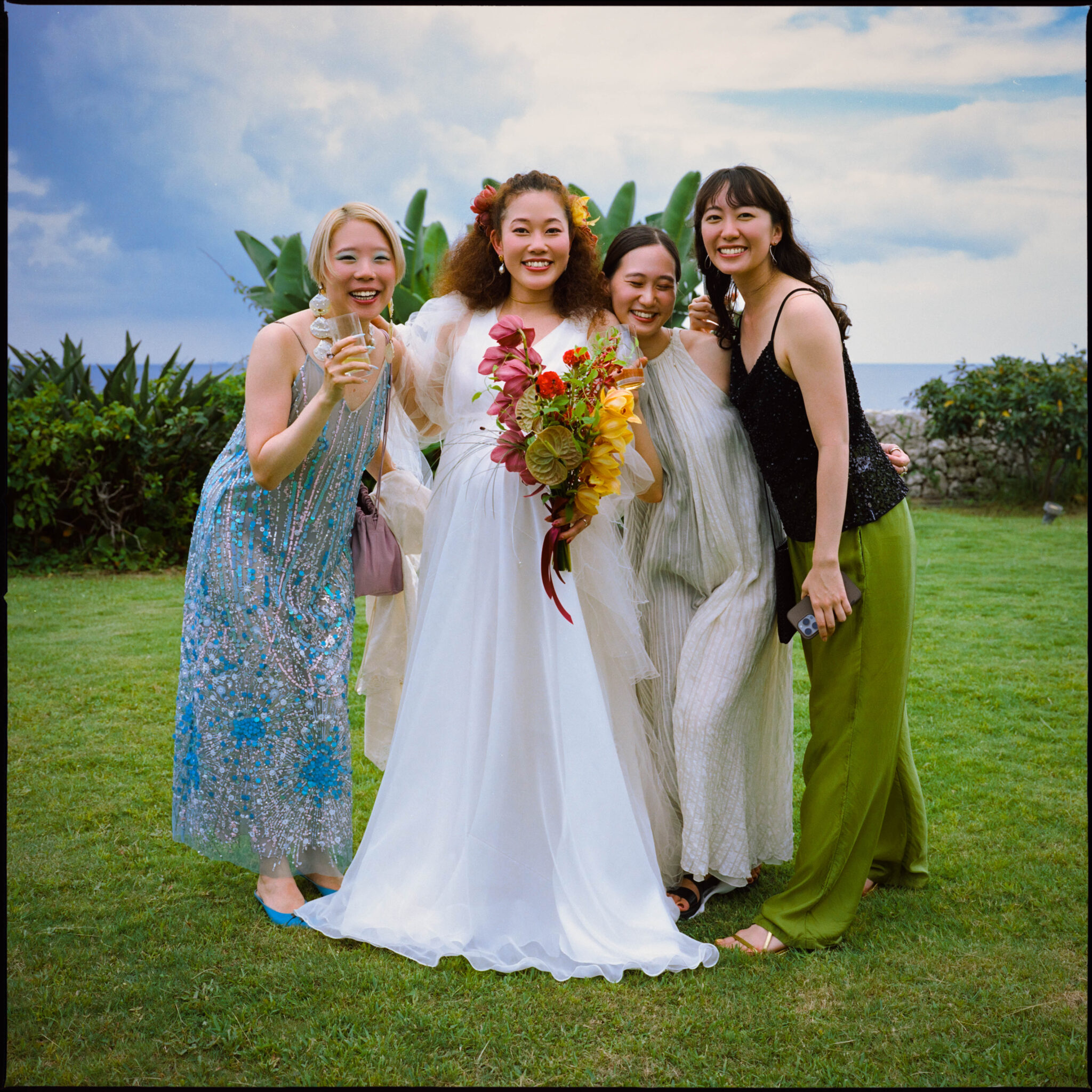 Bride with a tropical bouquet laughing with three friends on a seaside lawn, medium format film photograph