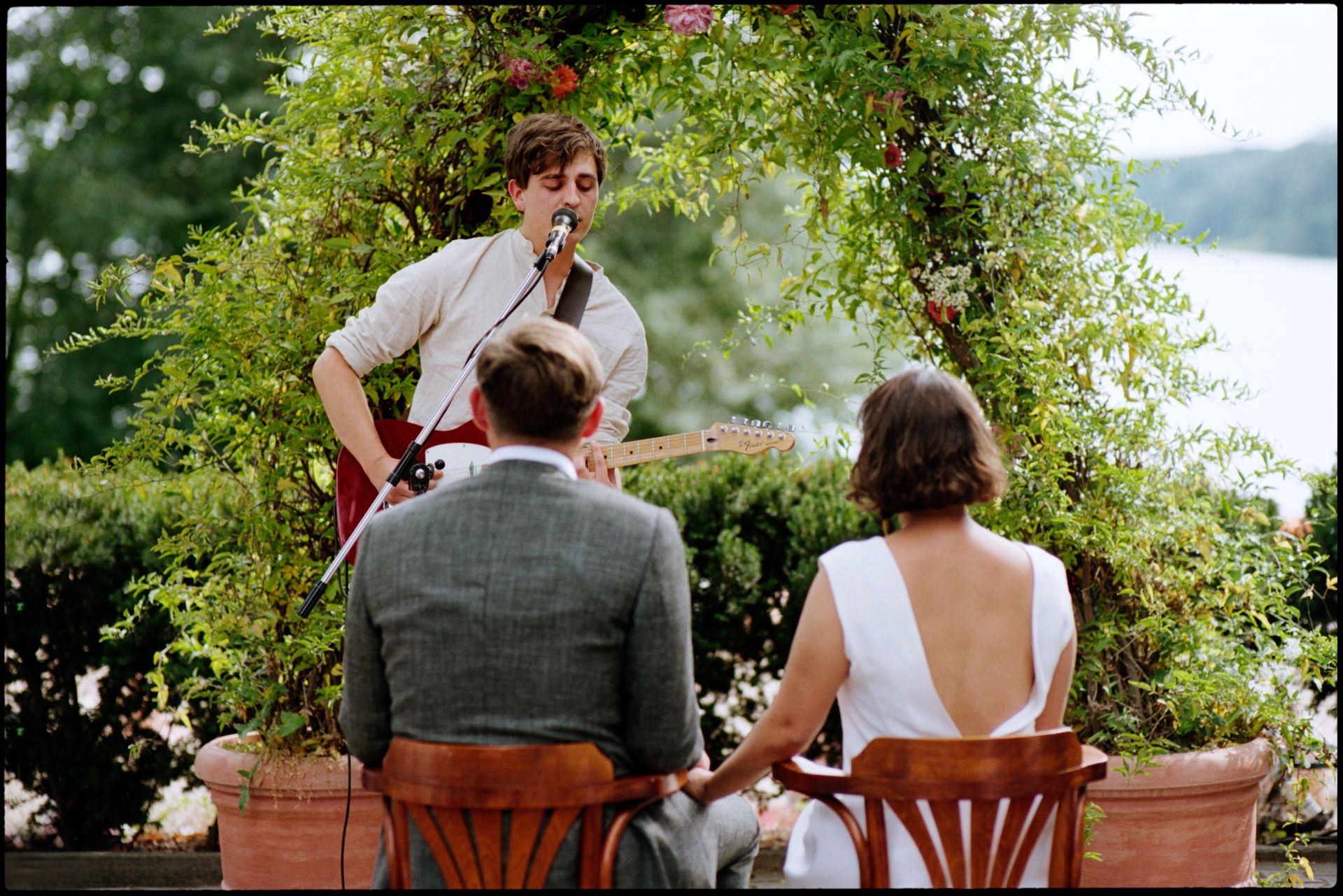 Musician playing guitar for the seated couple among greenery by the water, analogue film photograph