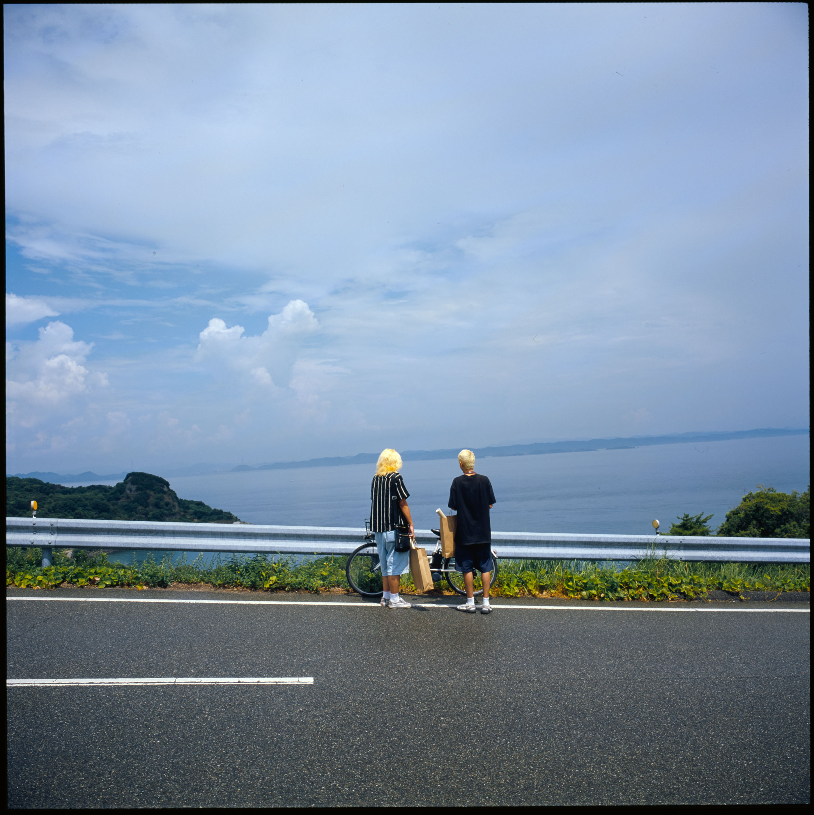 Two friends with paper shopping bags and a bicycle at a guardrail, looking out over the sea