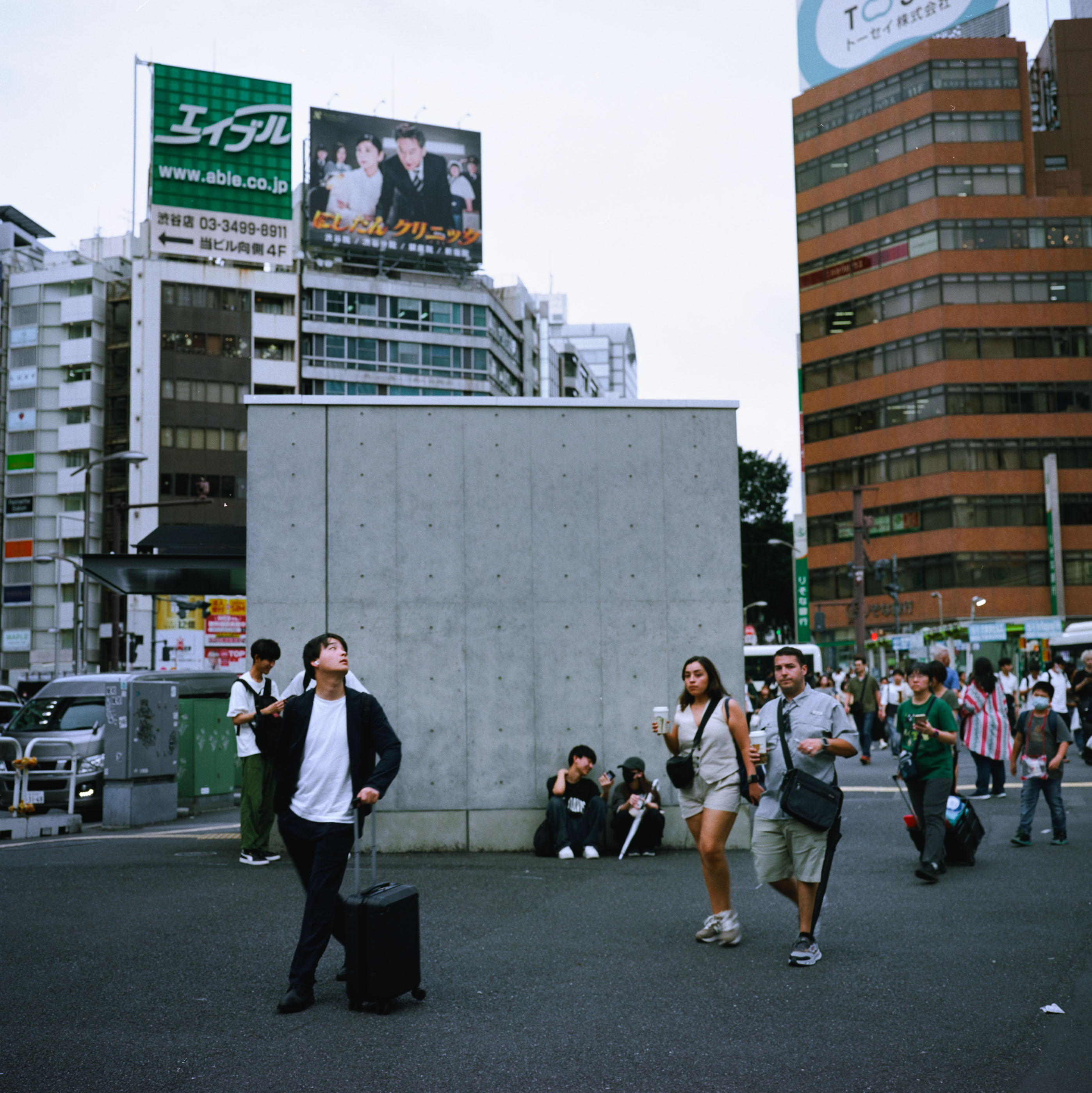 Traveller with a rolling suitcase looking up while crossing a busy square in Shibuya