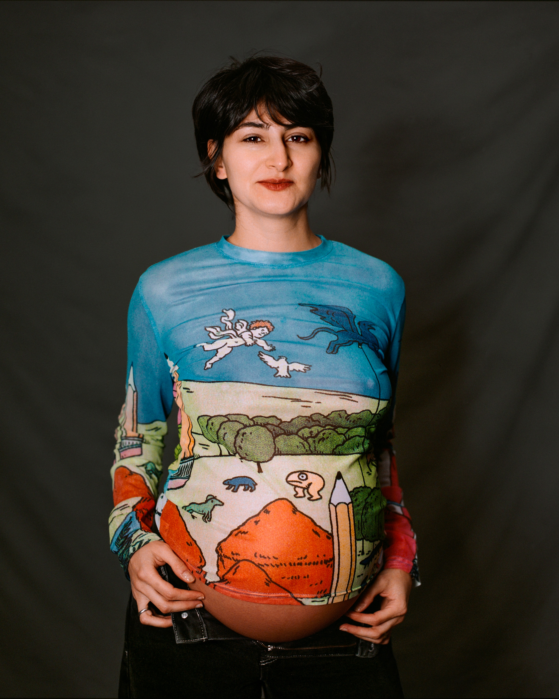 A pregnant woman cradling a painted vessel against a landscape-print top, analogue film portrait by Martina Manz, Berlin