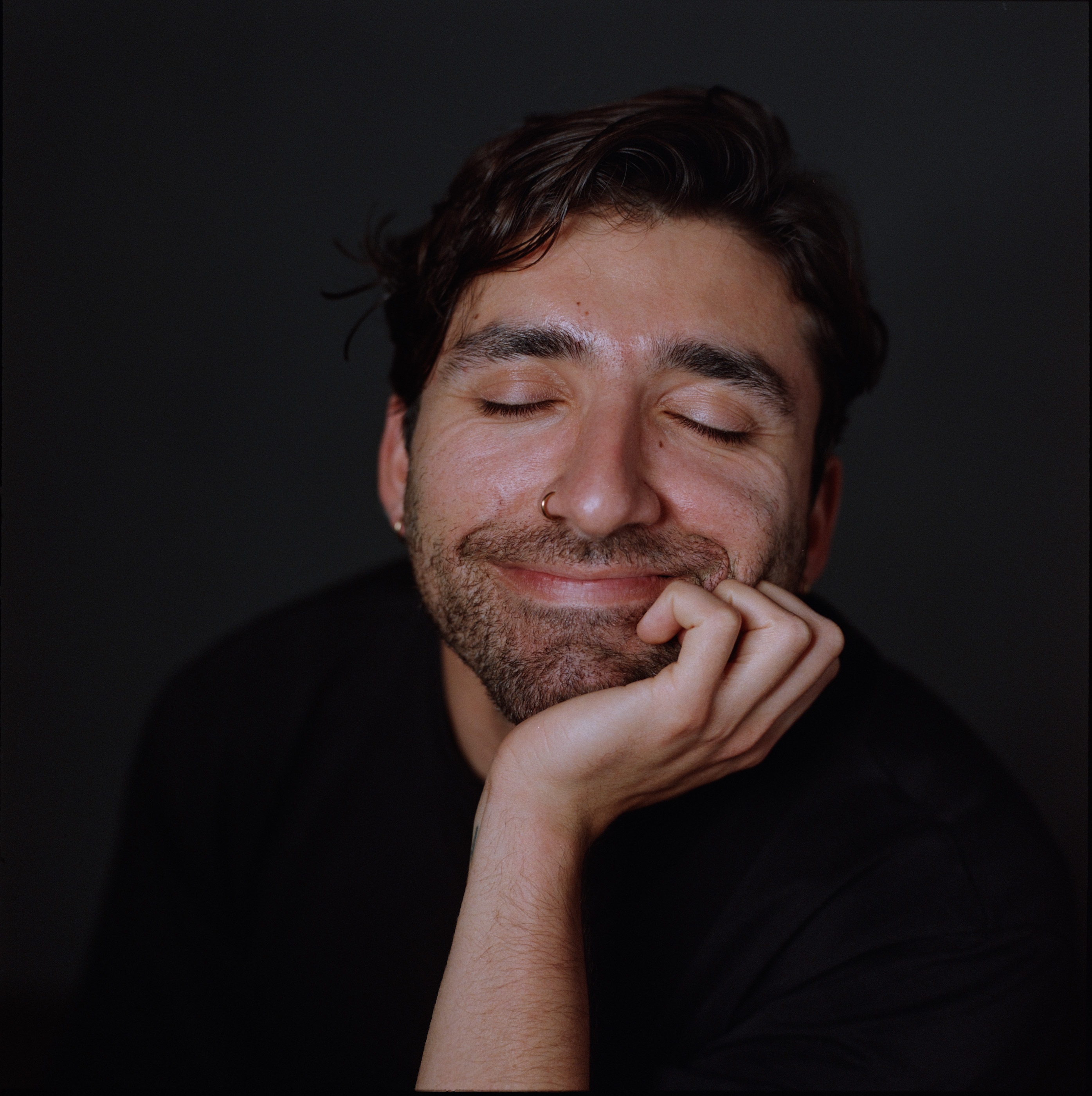 A man resting his chin on his hand with his eyes closed, against a dark backdrop, analogue film portrait by Martina Manz, Berlin