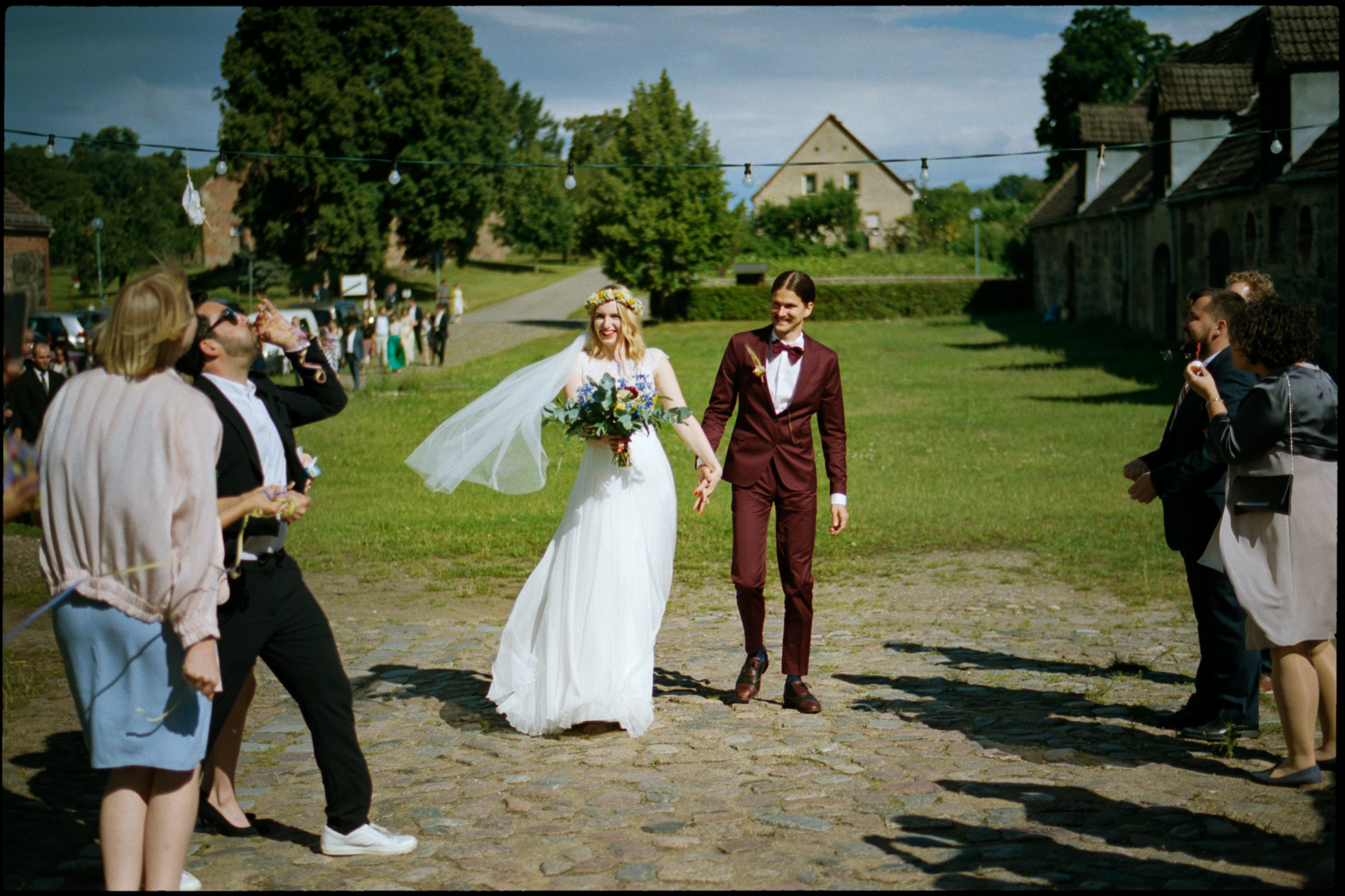Bride and groom walking hand in hand across a cobbled courtyard while guests blow soap bubbles, 35mm film photograph by Martina Manz